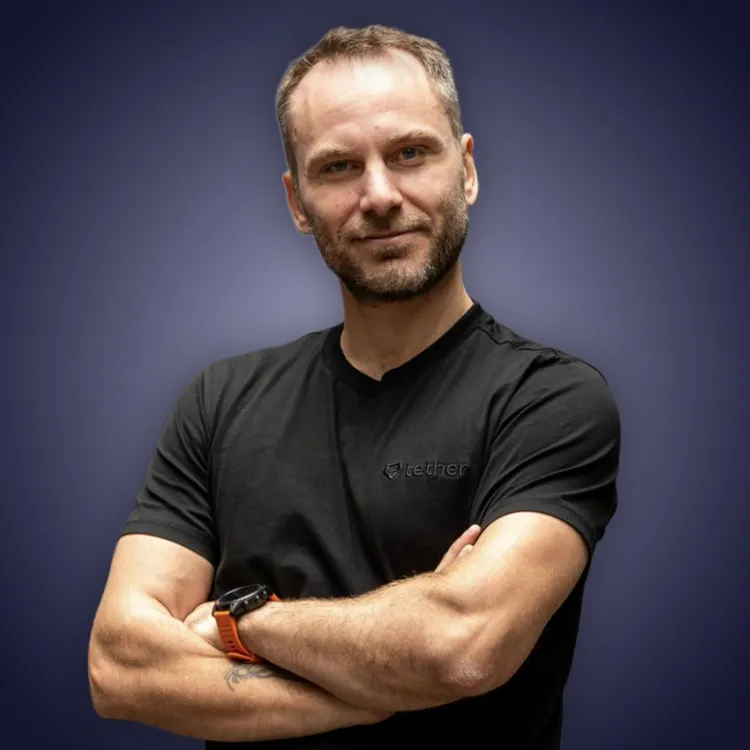 Man with crossed arms wearing a black Tether shirt standing confidently in front of a wall with Tether logos.
